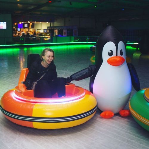A woman laughs while riding an inflatable bumper car shaped like a penguin in a brightly lit indoor rink.