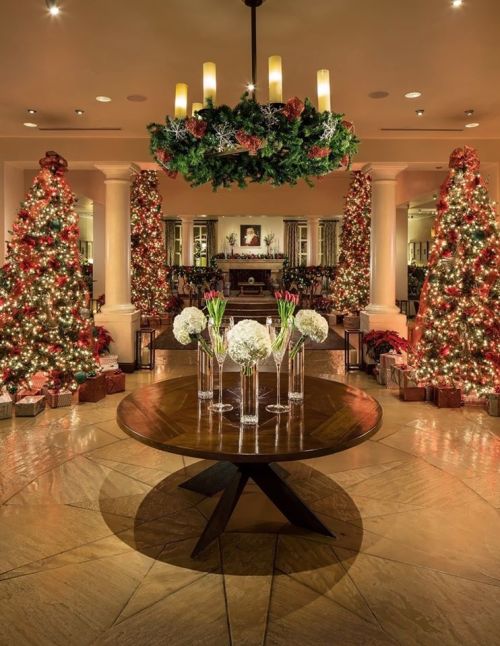 A festive lobby decorated with Christmas trees, gifts, and a floral centerpiece under a wreath and chandelier.