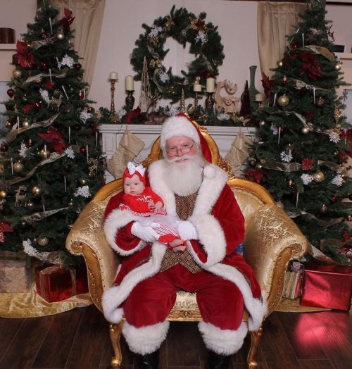 Santa Claus sits in a golden chair, holding a baby dressed in festive attire, surrounded by decorated Christmas trees.
