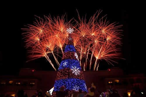 A brightly lit Christmas tree with colorful lights, surrounded by vibrant fireworks in the night sky.