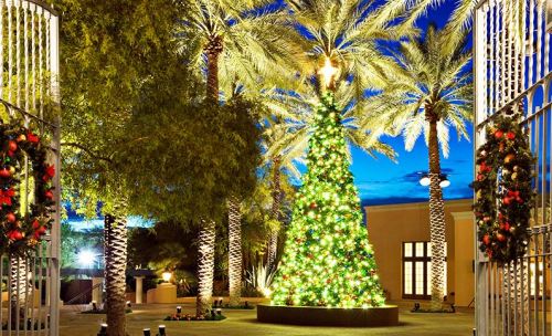 A beautifully lit Christmas tree surrounded by palm trees, with festive decorations, under a twilight sky.