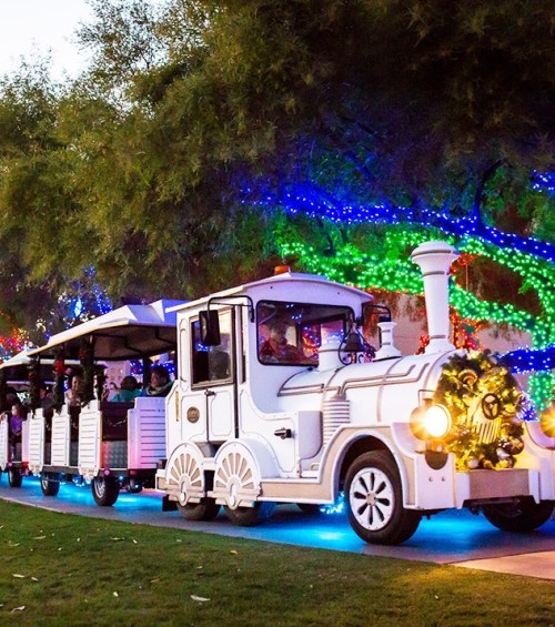 A festive train ride decorated with lights, carrying passengers through a park with colorful holiday decorations.