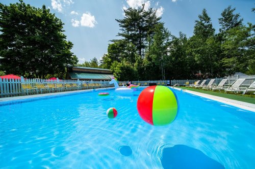 A sunny pool scene with colorful beach balls floating on the water, surrounded by lounge chairs and greenery.