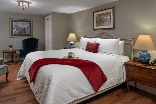 Cozy hotel room featuring a queen bed, bedside lamps, a chair, and artwork on the walls. Warm, inviting atmosphere.
