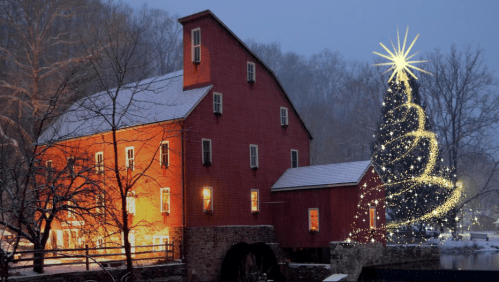 A red mill illuminated at night, surrounded by snow, with a sparkling Christmas tree nearby.