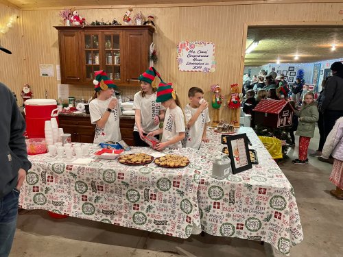 Children in elf hats serve cookies and drinks at a festive holiday event, with decorations and attendees in the background.