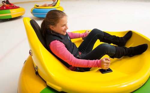 A girl smiles while riding in a yellow bumper car on an ice rink, with other colorful cars in the background.