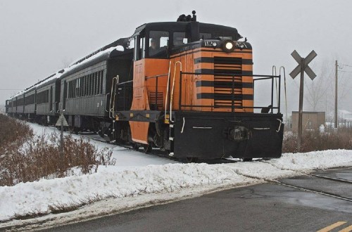 A vintage orange and black train engine pulls a passenger car through a snowy landscape on a foggy day.