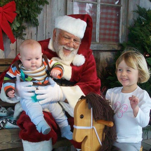 Santa Claus holds a baby while a young girl smiles beside them, with festive decorations in a cozy setting.
