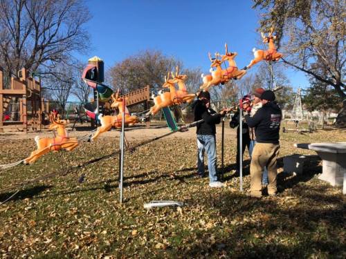 Four people adjust a row of orange reindeer decorations in a park with playground equipment in the background.