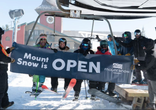 A group of skiers holds a banner reading "Mount Snow is OPEN" near a ski lift in a snowy landscape.