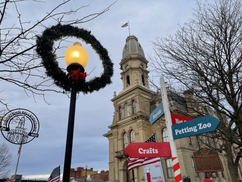 A festive street scene featuring a wreath on a lamp post, with a historic building and directional signs for local attractions.