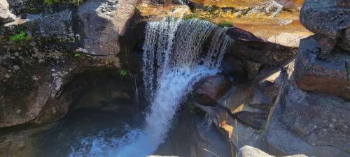 A cascading waterfall flows over rocky terrain, surrounded by lush greenery and sunlight.