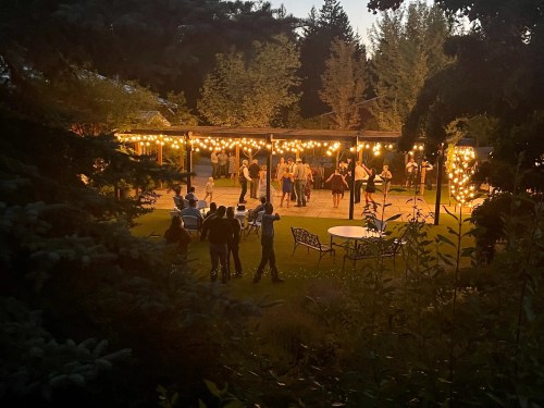 A lively outdoor gathering at dusk, with people dancing under string lights and surrounded by trees.