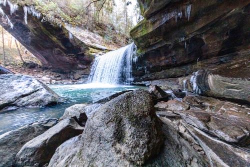 A serene waterfall cascades over rocks into a tranquil pool, surrounded by icy formations and lush greenery.