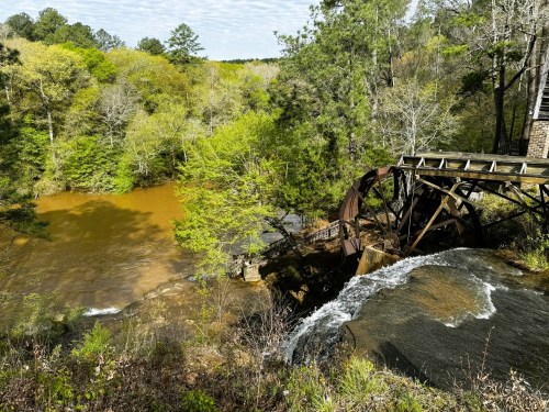 A scenic view of a river with a waterwheel, surrounded by lush green trees and a clear blue sky.
