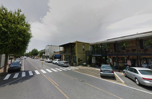 A street view of a small town with shops, parked cars, and a cloudy sky. Pedestrian crosswalk visible in the foreground.