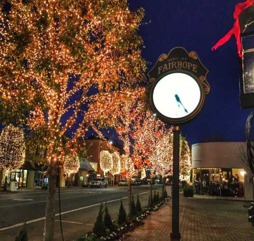 A festive street in Fairhope, adorned with twinkling lights on trees and a decorative sign at dusk.