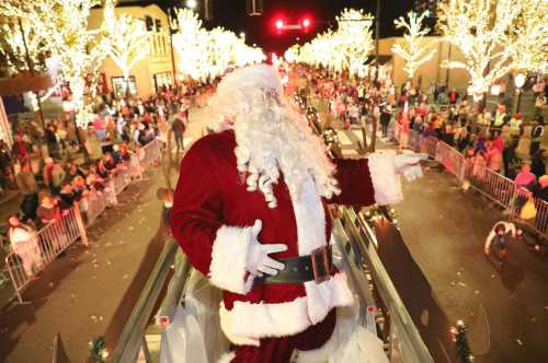 Santa Claus waves from a parade float, surrounded by festive lights and a cheering crowd on a lively street.