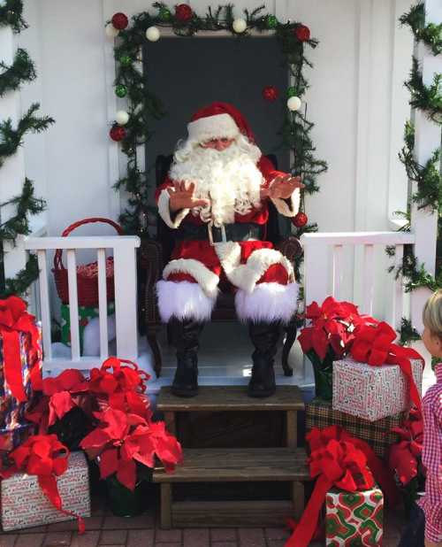 Santa Claus sits in a decorated booth surrounded by gifts and poinsettias, greeting a child.