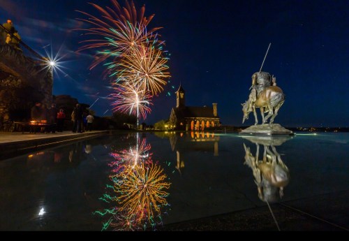 Colorful fireworks illuminate the night sky, reflecting in water near a statue and a quaint building.