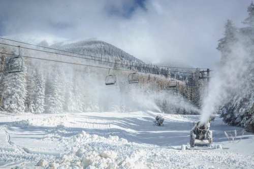 Snowmaking machines operate in a winter landscape, with ski lifts and snow-covered trees in the background.