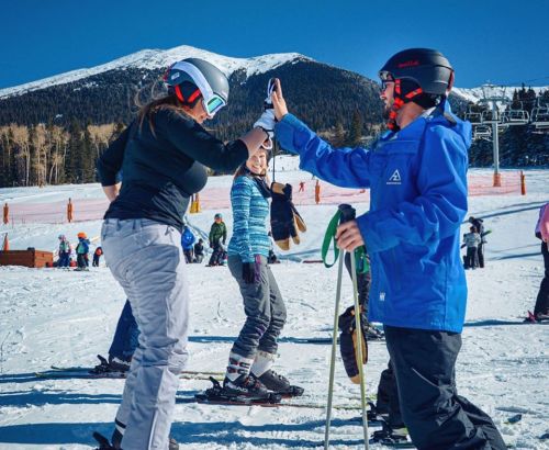 Two skiers high-five on a snowy slope, with others skiing in the background and mountains in the distance.