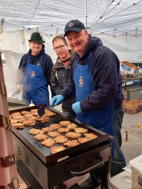 Three men in aprons cooking patties on a griddle at an outdoor event, smiling and engaging with the camera.