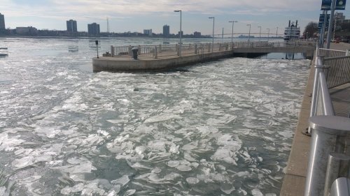 A frozen river with ice chunks near a waterfront dock and city skyline in the background on a cloudy day.