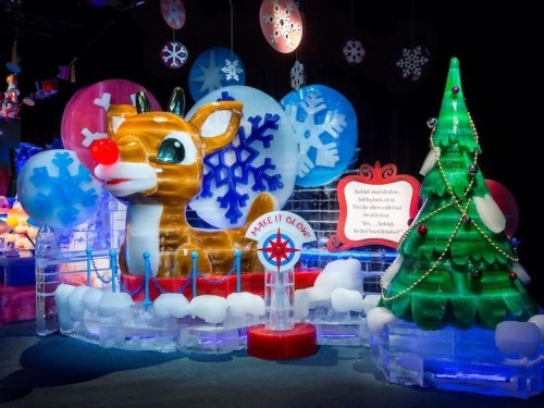 A colorful ice sculpture of Rudolph the Red-Nosed Reindeer beside a green Christmas tree and snowflakes.