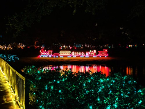 Colorful holiday lights illuminate a train display by a pond at night, reflecting on the water's surface.