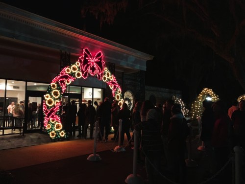 A festive entrance adorned with colorful Christmas lights and a large bow, with a line of people waiting outside.