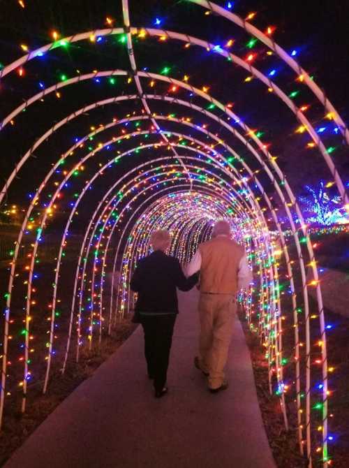 A couple walks hand-in-hand through a colorful, illuminated archway of holiday lights at night.