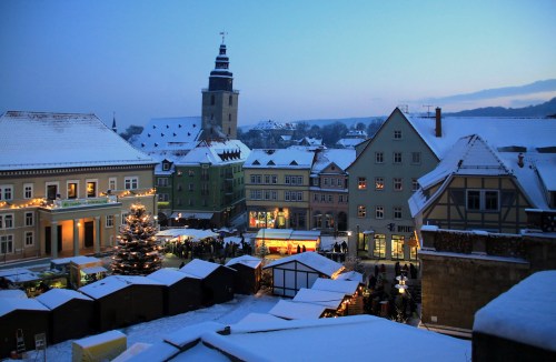 A snowy town square at dusk, featuring festive stalls, a Christmas tree, and a clock tower in the background.