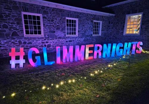 Colorful illuminated letters spell out "#GLIMMERNIGHTS" in a dark outdoor setting, with twinkling lights nearby.