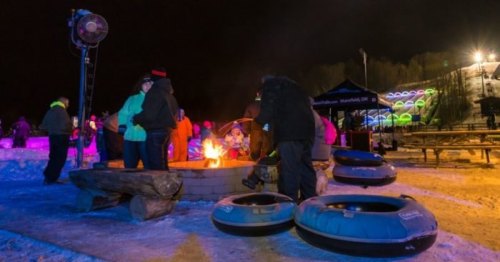 People gather around a fire pit at night, with snow tubing and colorful lights in the background.