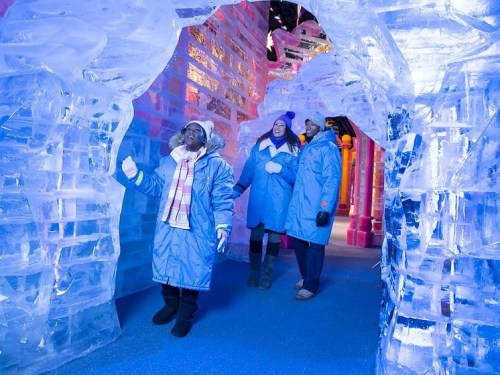 Three people in blue coats explore a colorful ice cave, surrounded by ice sculptures and illuminated by blue and pink lights.