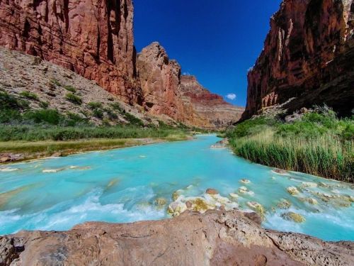 A vibrant turquoise river flows through a canyon with towering red rock formations under a clear blue sky.