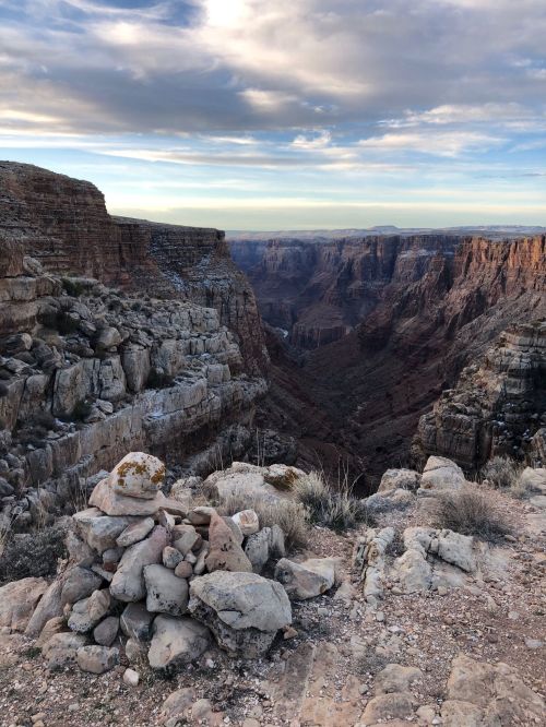 A rocky canyon landscape with layered cliffs and a vast valley under a cloudy sky.