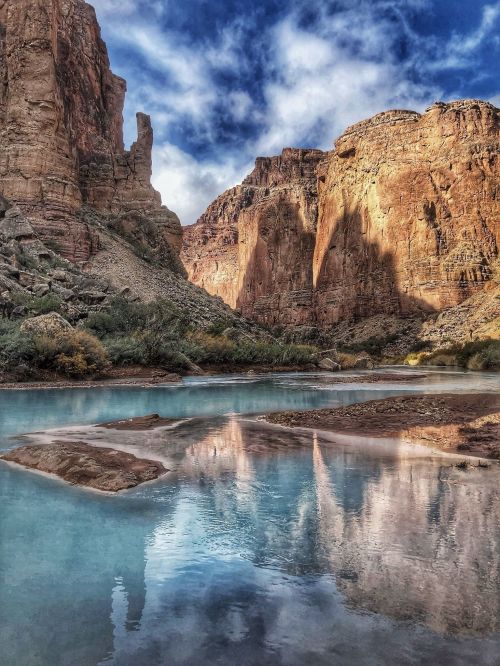 A serene landscape featuring towering rock formations reflected in a calm blue river under a cloudy sky.