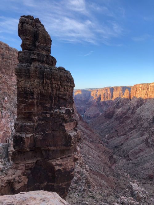 A towering rock formation stands against a backdrop of the Grand Canyon, illuminated by the soft glow of sunset.