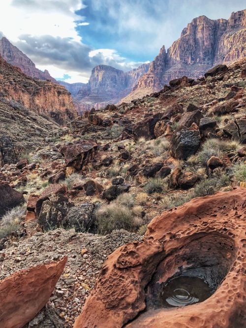 A rocky landscape with mountains in the background and a small pool of water in the foreground under a cloudy sky.