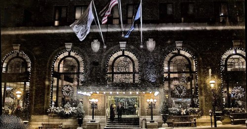A historic hotel entrance adorned with holiday decorations and snow, featuring large arched windows and flags.