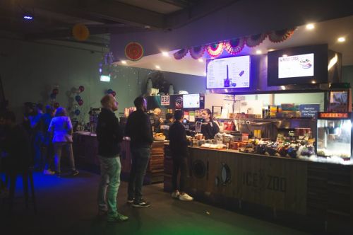 A busy food counter in a dimly lit venue, with customers ordering and colorful decorations in the background.