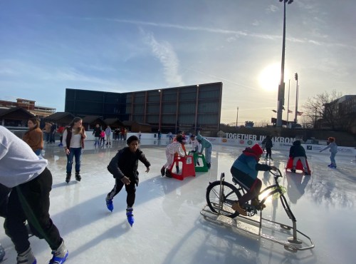 A busy outdoor ice skating rink with people skating, using aids, and a bike on the ice under a sunny sky.