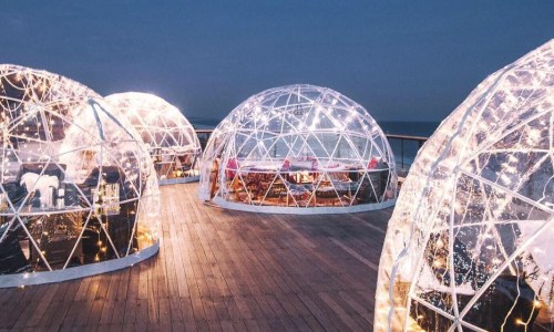 A scenic outdoor dining area featuring illuminated glass igloos on a wooden deck by the ocean at dusk.