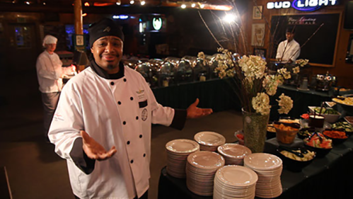 A chef in a white uniform stands with open arms in a buffet setting, showcasing a variety of dishes and plates.