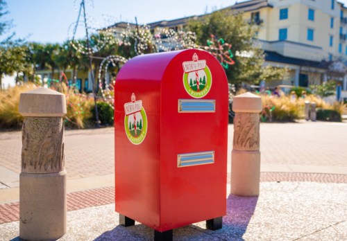 Bright red mailbox with festive decorations, set in a landscaped area with buildings in the background.