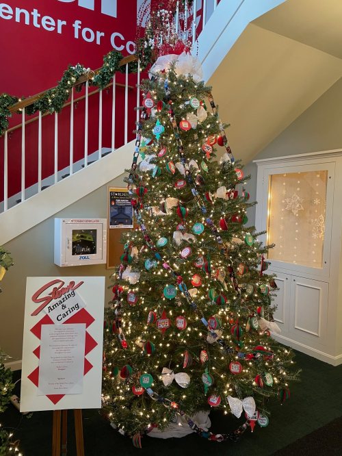A decorated Christmas tree with colorful ornaments and lights, next to a sign in a festive indoor setting.