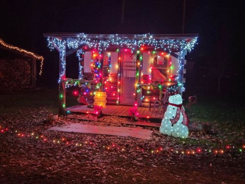 A cozy cabin decorated with colorful holiday lights and a snowman in the foreground, set in a dark, festive night.